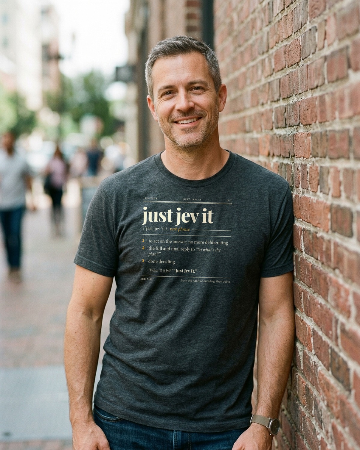 Man smiling against a brick wall in a charcoal tee with the Just Jev It dictionary entry on the chest